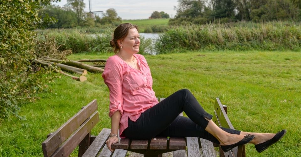Ontspannen vrouw buiten aan het water met een lach op haar gezicht – symbool voor balans, plezier en werkgeluk vanuit eigen ervaring.