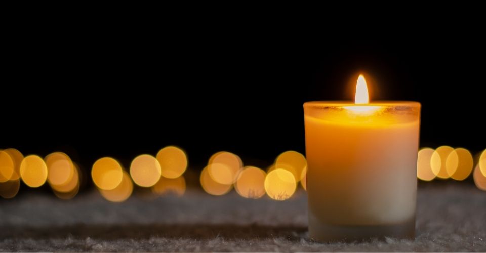 Brandende kaars in het donker met zachte lichtjes op de achtergrond – een symbool van herinnering, afscheid en verbondenheid.