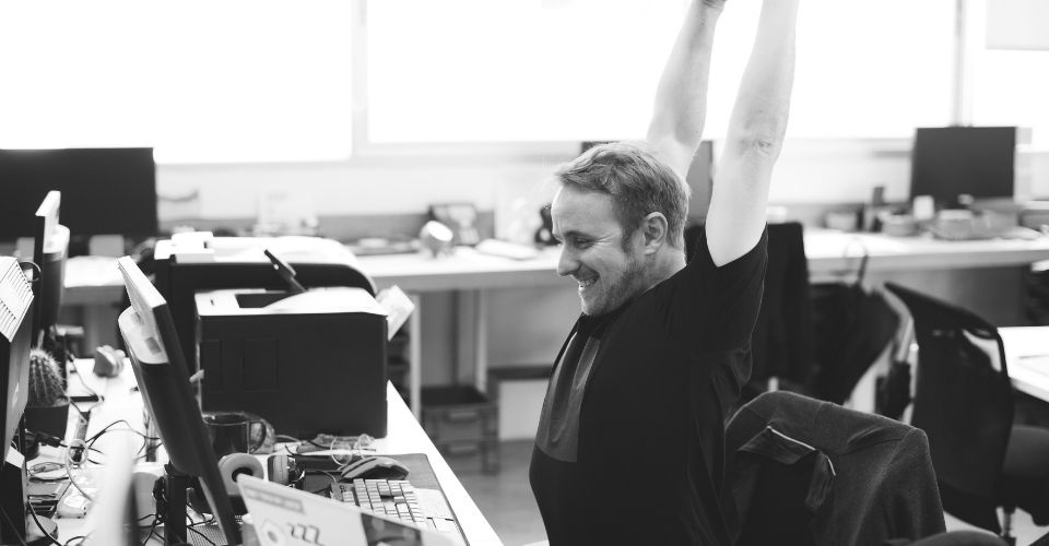 Man rekt zich uit met een glimlach achter zijn bureau – een moment van ontspanning tijdens een werkdag als voorbeeld van omgaan met werkstress.