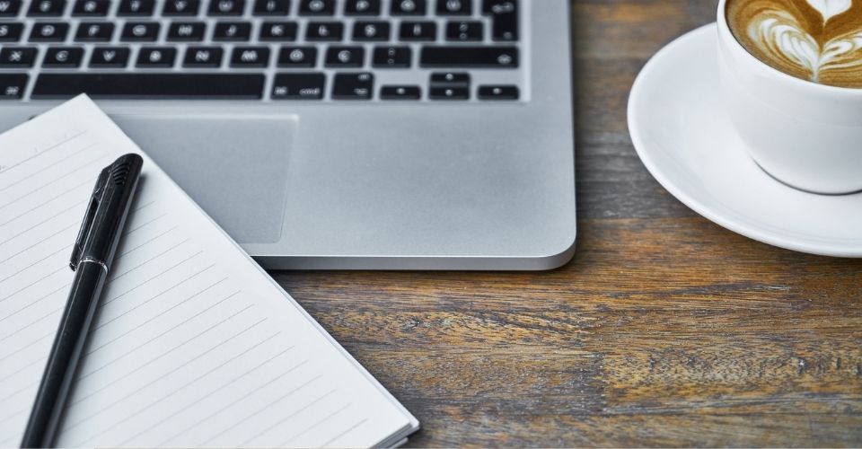 Notitieboek, pen, laptop en cappuccino op houten tafel – een uitnodigende setting om gedachten op papier te zetten en tot rust te komen.