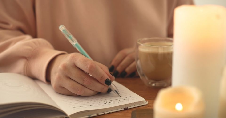 Vrouw schrijft in haar dagboek bij kaarslicht, met een kop warme koffie - een dagelijkse gewoonte om de dag positief af te sluiten en gevoelens om te buigen.