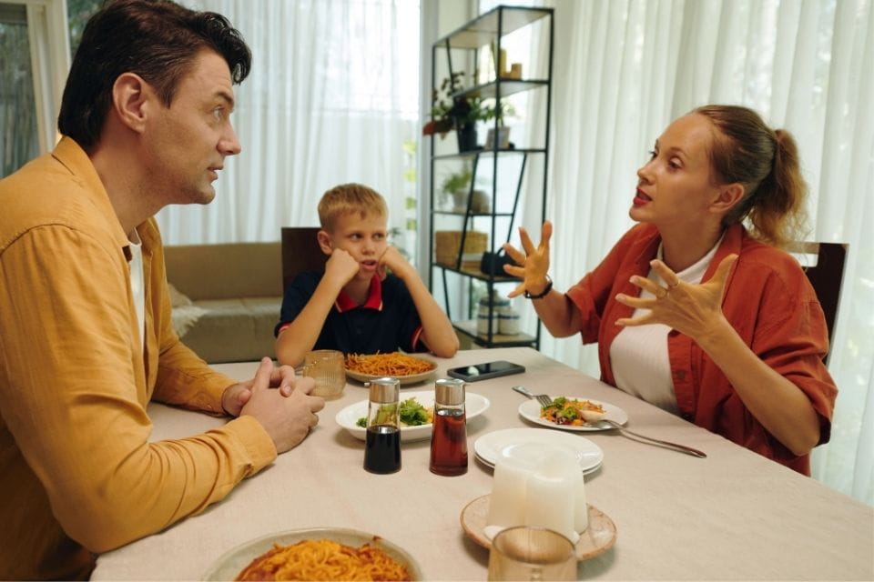 Ouders in gesprek tijdens het avondeten met hun kind erbij, zichtbaar spanning en miscommunicatie in de lucht.